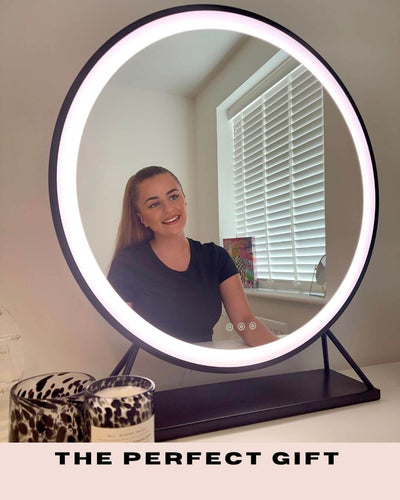Woman sitting at dressing table with Grace Round Hollywood Mirror in black, showcasing customizable LED lighting for beauty enthusiasts.