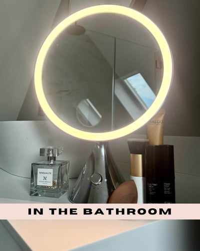 LumiGlam Pro Cosmetic Vanity Mirror in chrome with LED light on bathroom counter, surrounded by perfume and makeup products.