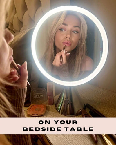 Woman using LumiGlam Pro vanity mirror with LED lighting for detailed makeup application on bedside table.