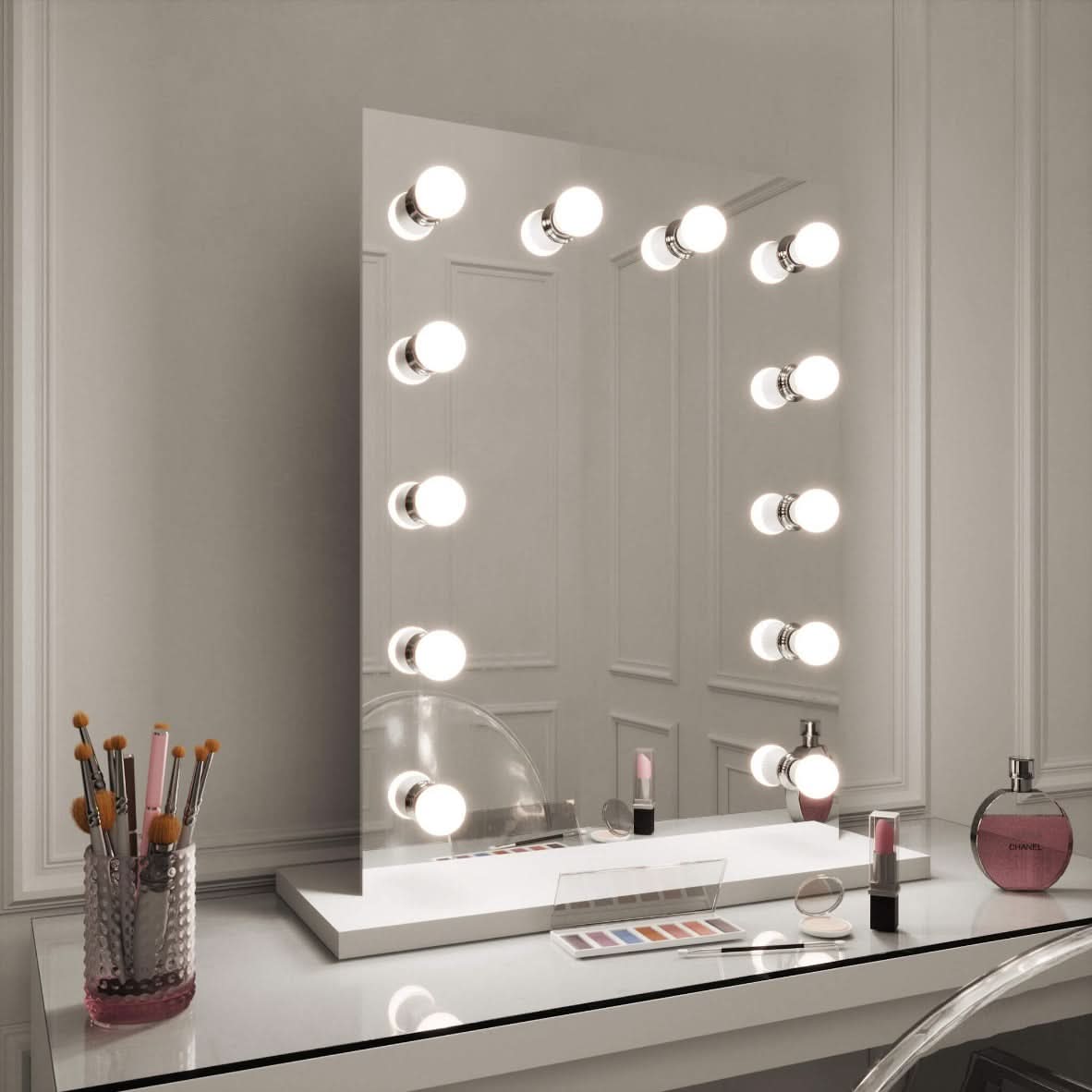 The hollywood mirrors Alicia Hollywood Mirror 60cmx80cm features twelve LED bulbs, a dimmer switch, and comes with a transparent chair. On the table: makeup brushes in a clear holder, eyeshadow palette, lipstick, and perfume bottle.