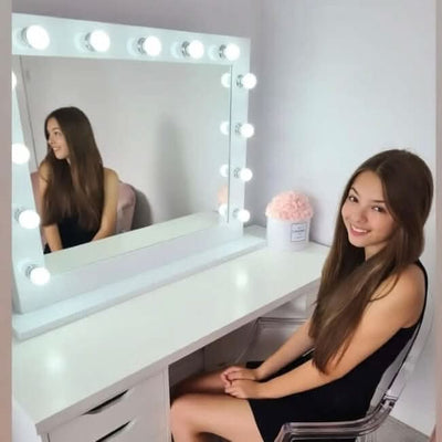 A young woman with long brown hair smiles at the camera while sitting at a white vanity fitted with the hollywood mirrors Audrey Hollywood Mirror White 100x80cm. A pink bouquet sits on the table in a bright, minimalist room.