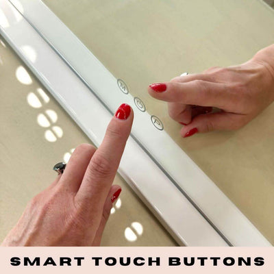 A person with red nail polish touches smart touch buttons on a white Audrey Hollywood Mirror 100x80cm by hollywood mirrors, set beneath glowing studio lighting, with the text SMART TOUCH BUTTONS at the bottom.