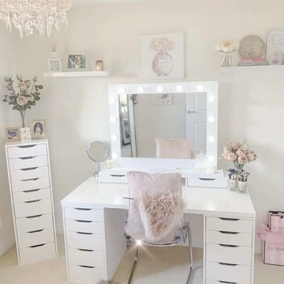 A softly decorated room with a bright white vanity, the Audrey Hollywood Mirror White 100x80cm by hollywood mirrors featuring tri-colour LED bulbs, a white desk with drawers, clear chair with pink pillow, flowers, and decor on shelves and tabletop.