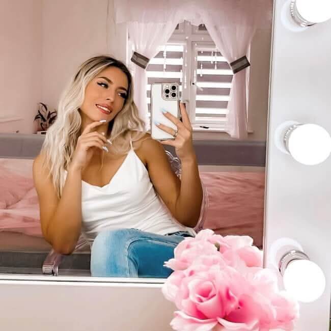 A woman with long blonde hair wearing a white top and blue jeans takes a mirror selfie in a bright room. Pink flowers decorate the scene, and the Audrey Hollywood Mirror White 100x80cm by hollywood mirrors is visible along the edge.