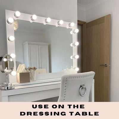 The Audrey Hollywood Mirror White 100x80cm by hollywood mirrors sits on a white dressing table with tri-colour LED bulbs, a cushioned chair, makeup brushes, tissues, and a round mirror. Text below reads USE ON THE DRESSING TABLE.