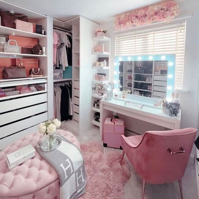 A chic dressing room features open shelves for handbags, shoes, and clothes, a vanity with the Audrey Hollywood Mirror White 100x80cm by Hollywood Mirrors, studio lighting, a pink chair, fluffy pink rug, and a pink ottoman topped with flowers and a book.