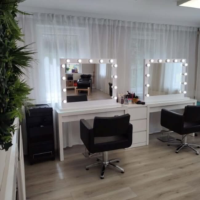 Two black salon chairs face white vanities with Audrey Hollywood Mirror White 100x80cm by hollywood mirrors, featuring Tri-Colour LED bulbs that brighten the room with sheer curtains, light wood floors, green wall plants, and neatly arranged beauty products.