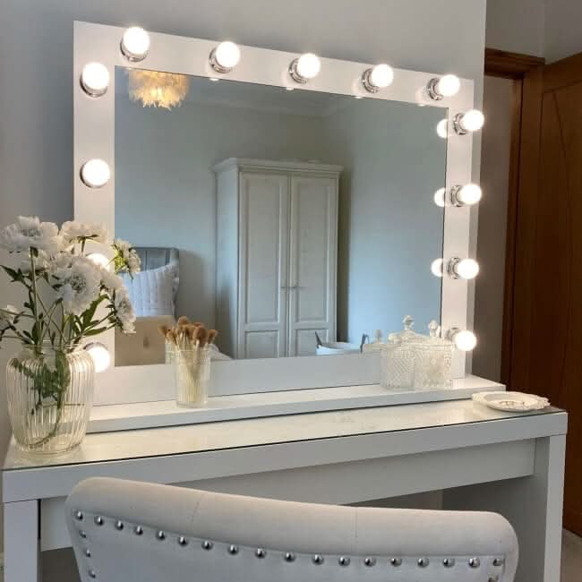 The Audrey Hollywood Mirror White 100x80cm by hollywood mirrors features studio lighting with round bulbs. A cushioned chair with silver studs, white flowers, makeup brushes, and glass containers complete the look; a wardrobe is reflected in the mirror.