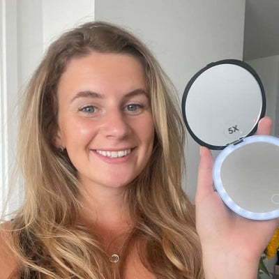 A woman with long blonde hair smiles at the camera while holding up an open Black Compact Mirror with Light, its round edge softly illuminated.