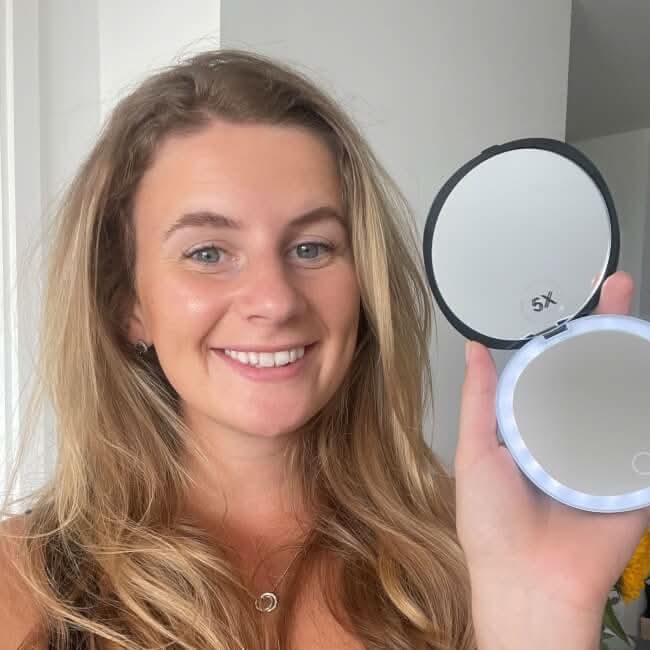 The Black Compact Mirror with Light is a round, open LED-lighted makeup mirror with a touch-sensitive power button, perfect for easy use anywhere. It sits on a glossy pink surface and is portable for your convenience.