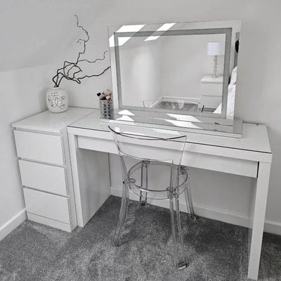 A modern vanity setup with the hollywood mirrors Hollywood Crystal Pro Mirror 80x60cm, featuring dimmable lights, a clear acrylic chair, small white drawers, minimalist decor with a vase and white piece, plus grey carpet and white walls.
