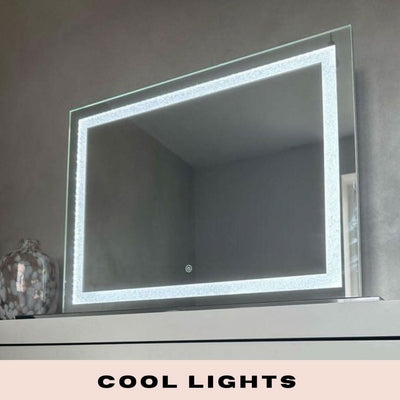 The Hollywood Crystal Pro Mirror 80x60cm by hollywood mirrors features illuminated LED strips around its border, sits on a white surface in a modern gray room, and offers dimmable cool lighting. A patterned glass vase is placed to the left of the mirror.