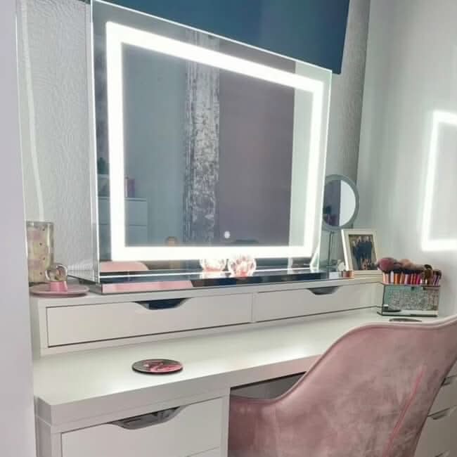 A white vanity desk with drawers, a hollywood mirrors Hollywood Crystal Pro Mirror 80x60cm framed by dimmable LED strips, a pink velvet chair, makeup brushes, a framed photo, and decorative decor on the tabletop.
