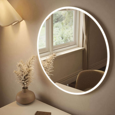 Round backlit mirror with ambient lighting on a wall above a desk, featuring a window reflection and decorative vase.