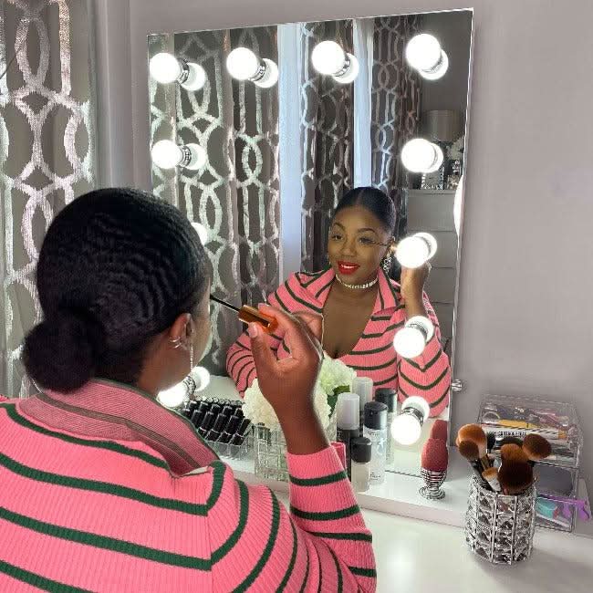 The hollywood mirrors Alicia Hollywood Mirror 60cmx80cm features twelve LED bulbs, a dimmer switch, and comes with a transparent chair. On the table: makeup brushes in a clear holder, eyeshadow palette, lipstick, and perfume bottle.