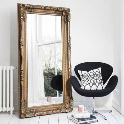 Charles Leaner Mirror in Gold with ornate carved detailing, elegantly placed beside a modern chair in a stylish room setting.