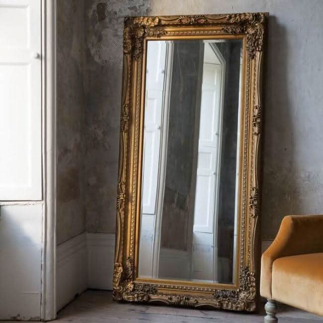 Charles Leaner Mirror in gold with carved detailing, wood and resin frame, styled against a rustic wall next to a mustard chair.