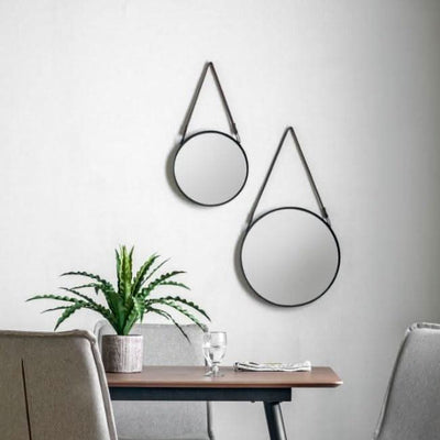 Set of two black metal framed Marston hanging mirrors with faux leather straps on a wall above a modern dining table and plant.