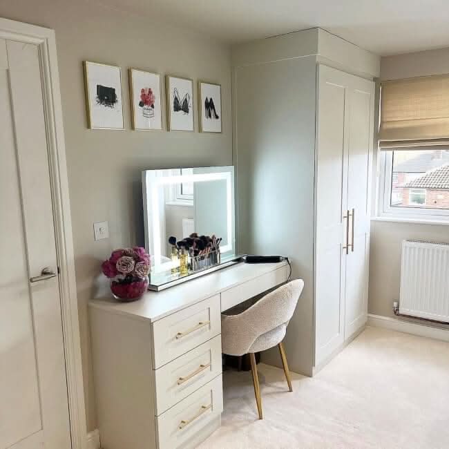 A modern vanity with drawers, the hollywood mirrors Hollywood Crystal Pro Mirror 80x60cm with dimmable lights, makeup brushes, and flowers sits by a white wardrobe and a window with beige blinds. Four art prints are above the vanity.