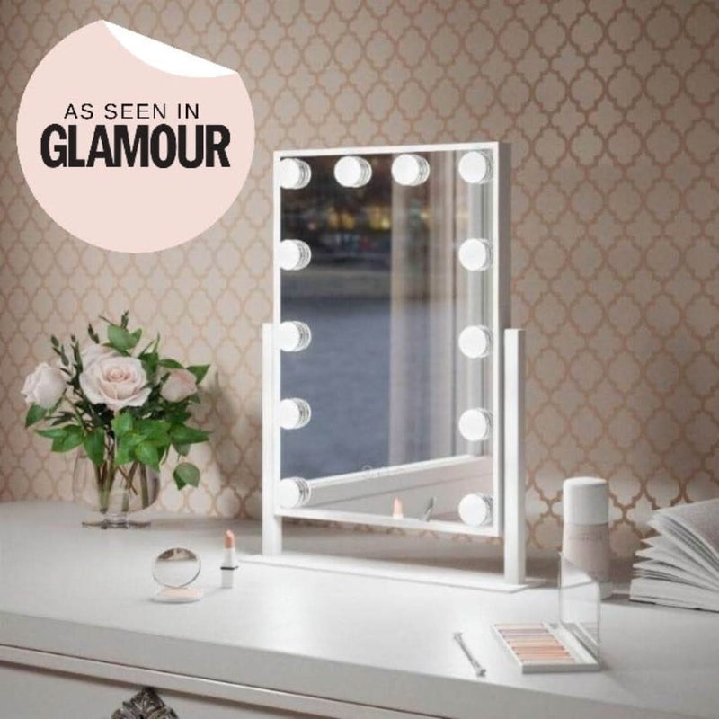 The Olsen Hollywood Mirror White 47x37cm by hollywood mirrors sits on a white vanity with adjustable LED bulbs, makeup items, a lipstick, flowers in a vase, and an open book against patterned wallpaper.