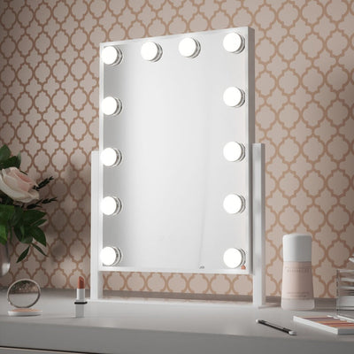 The Olsen Hollywood Mirror White 47x37cm by hollywood mirrors, featuring LED bulbs and adjustable brightness, sits on a desk with patterned wallpaper, alongside a flower vase, lipstick, jar, crystal ball, and closed makeup container.