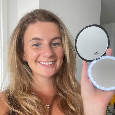 A woman with long blonde hair smiles at the camera while holding up an open Black Compact Mirror with Light, its round edge softly illuminated.