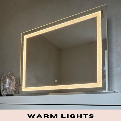 The Hollywood Crystal Pro Mirror 80x60cm by hollywood mirrors features a built-in warm LED strips border and is shown wall-mounted above a white surface with a decorative vase. WARM LIGHTS appears at the bottom of the image.