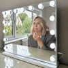 A woman with blond hair applies lipstick while looking into a Jessica Hollywood Mirror 58cmX46cm, its versatile LED lighting brightening the room framed by lattice windows.