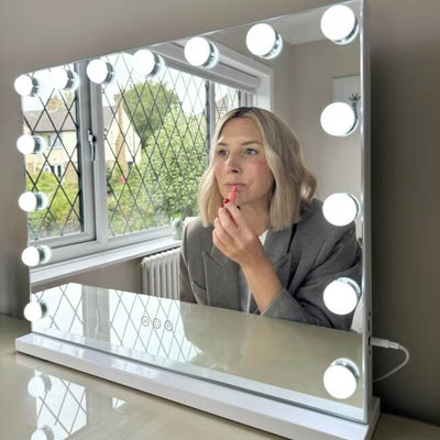 A woman with blond hair applies lipstick while looking into a Jessica Hollywood Mirror 58cmX46cm, its versatile LED lighting brightening the room framed by lattice windows.