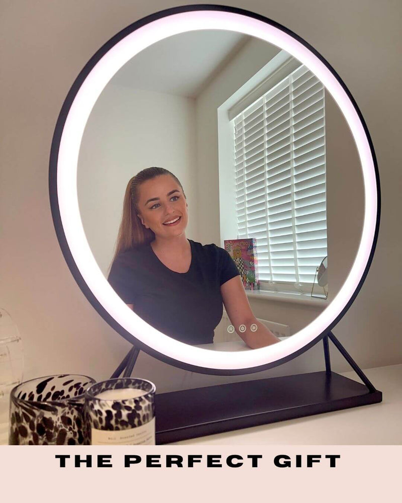 Round black frame Hollywood mirror with customizable LED lighting on a white dressing table next to a 'Glow Up' neon sign.