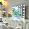 The Jessica Hollywood Mirror 58cmX46cm with LED bulbs offers versatile lighting by the window. A fluffy chair, storage drawers, and shelves for plants and decor complement the setup, while a green lamp adds a glow beside the mirror.