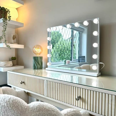 The Jessica Hollywood Mirror 58cmX46cm with LED bulbs offers versatile lighting by the window. A fluffy chair, storage drawers, and shelves for plants and decor complement the setup, while a green lamp adds a glow beside the mirror.