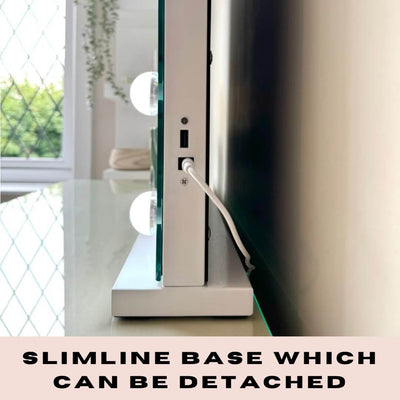 A close-up side view of the Jessica Hollywood Mirror 58cmX46cm with built-in LED bulbs on a slim, detachable base, placed on a reflective surface. A connected power cable and a window in the background are visible. Text: Slimline base can be detached.