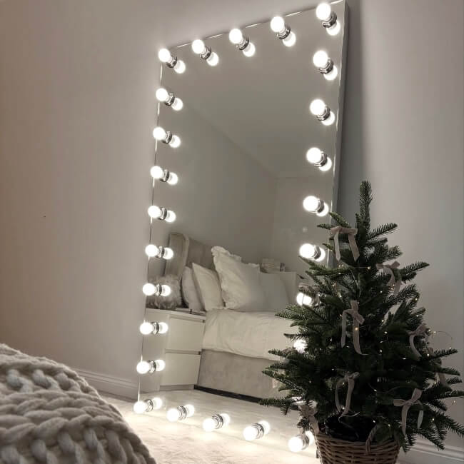 A cozy bedroom with a large, illuminated mirror bordered by light bulbs, reflecting a neatly made bed with white bedding. A small decorated Christmas tree stands on the floor nearby.