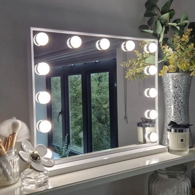 A Margot Small Hollywood Mirror 50x42cm with round bulb lights is displayed on a shelf beside a vase of green foliage, a candle, and flower-shaped decor, reflecting the window and trees outside.