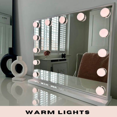 A Margot Small Hollywood Mirror 50x42cm with warm, round lights and adjustable brightness sits on a white desk. Decorative objects and a chair appear in the reflection. The text below reads: WARM LIGHTS.