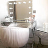 A stylish vanity setup includes the Penelope Hollywood Mirror Wall Mounted 80cmx60cm with LED bulbs, a mirrored desk for makeup essentials, a plush white chair, and a matching side table topped with white flowers—ideal for any makeup artist.