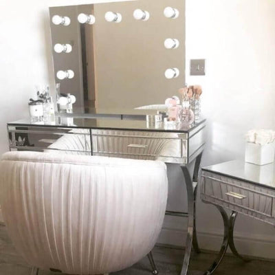 A stylish vanity setup includes the Penelope Hollywood Mirror Wall Mounted 80cmx60cm with LED bulbs, a mirrored desk for makeup essentials, a plush white chair, and a matching side table topped with white flowers—ideal for any makeup artist.