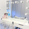 A white vanity table with the Penelope Hollywood Mirror Wall Mounted 80cmx60cm featuring LED bulbs, makeup brushes in holders, and a chair draped with a fluffy gray throw—ideal for any aspiring makeup artist. Closed wardrobe doors in the background.