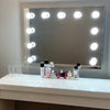 The Penelope Hollywood Mirror Wall Mounted 80cmx60cm is a large vanity mirror with ten LED bulbs, set above a clean, modern table holding two empty glasses and several bottles of perfume and beauty products—ideal for makeup artists.