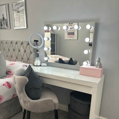 The Penelope Hollywood Mirror Wall Mounted 80cmx60cm, with LED bulbs, hangs on a gray wall above a modern vanity table. Nearby are a tufted chair and pink lips bedding—ideal for aspiring makeup artists with chic decor accents.