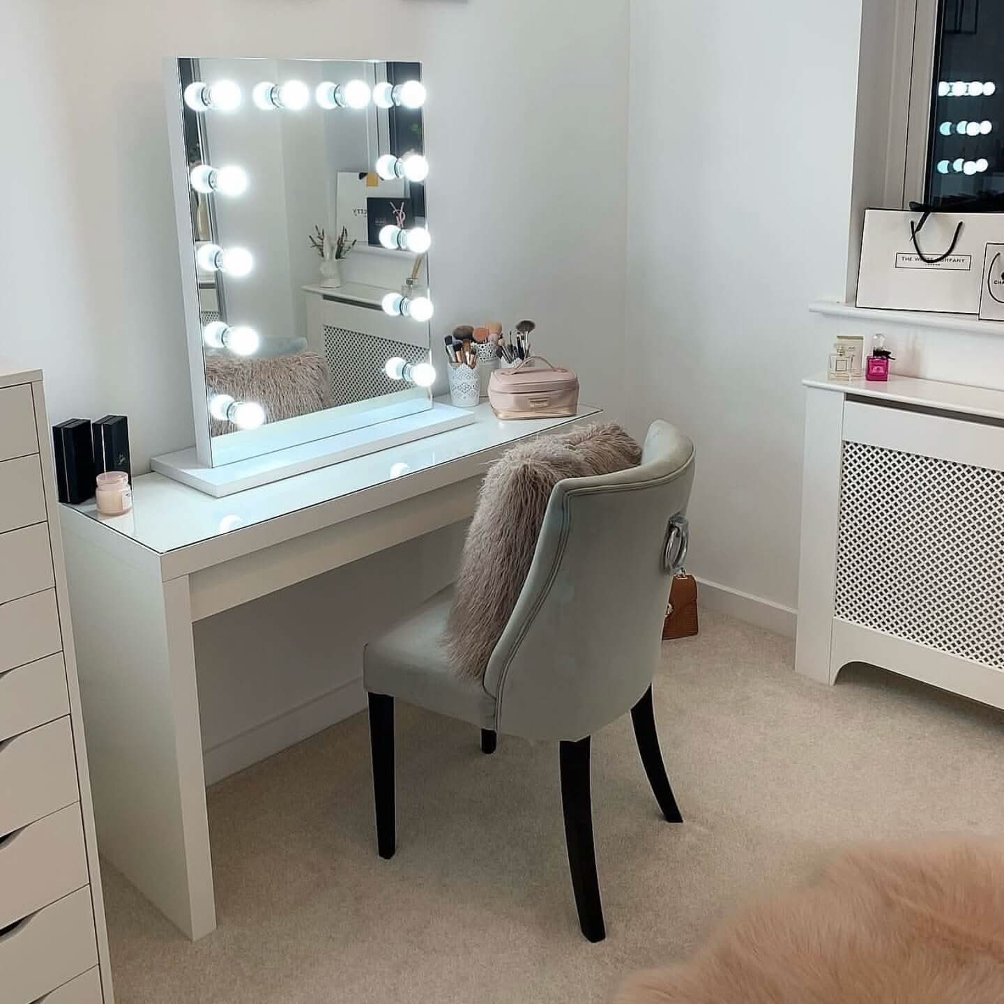 The hollywood mirrors Alicia Hollywood Mirror 60cmx80cm, framed by LED bulbs with a dimmer switch, stands on a vanity table holding makeup and brushes. A plush chair with a furry pillow adds comfort in the tidy, well-lit room.