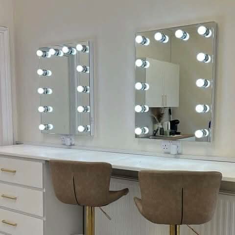 Two brown upholstered chairs sit before a white vanity table with drawers. Above it, two hollywood mirrors Alicia Hollywood Mirror 60cmx80cm, each with LED bulbs and dimmer control, are mounted on the wall.