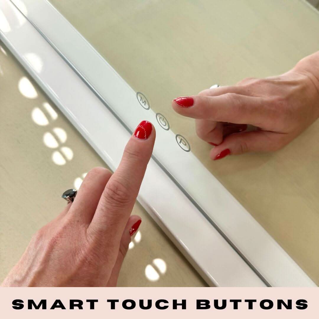 A person with red nail polish touches smart touch buttons on a white Audrey Hollywood Mirror 100x80cm by hollywood mirrors, set beneath glowing studio lighting, with the text SMART TOUCH BUTTONS at the bottom.
