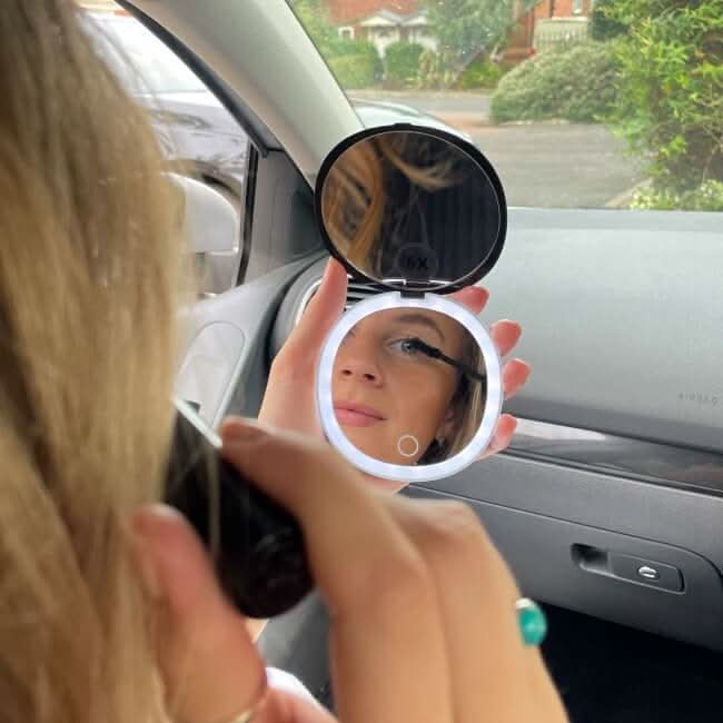 Using the Black Compact Mirror with Light, a person applies mascara in their car, clearly seeing their reflection in the magnified mirror as LED lights illuminate their face. The car’s dashboard and window are visible in the background.