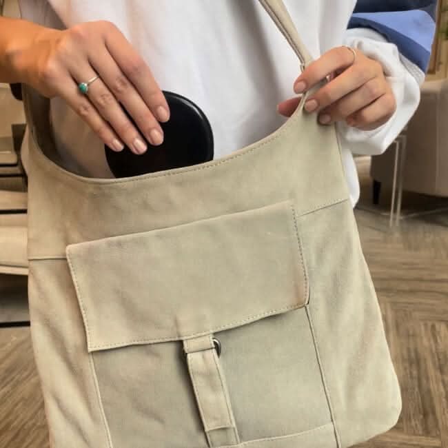 A person wearing a white long-sleeve shirt is placing a Black Compact Mirror with Light into a beige suede shoulder bag featuring a front pocket and buckle detail.