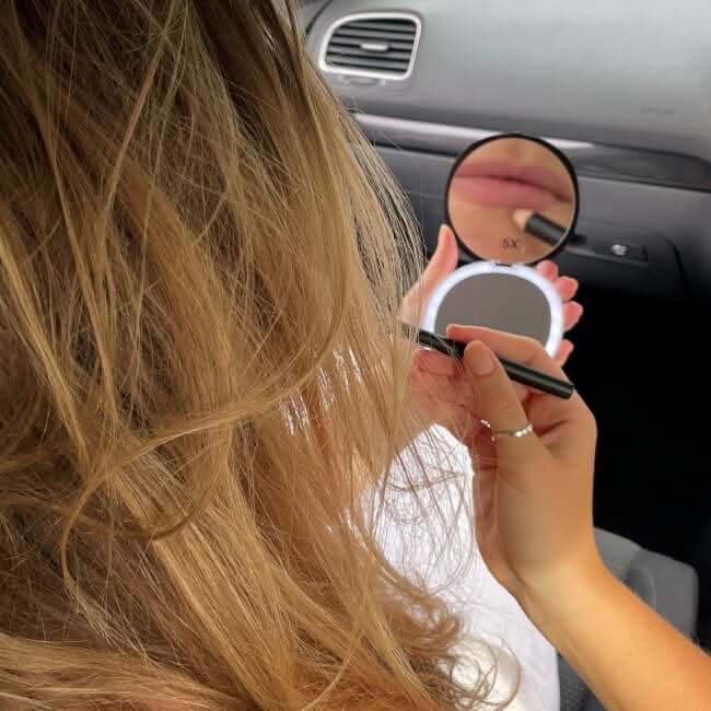 A woman with long blonde hair sits in a car, applying lipstick while holding the Black Compact Mirror with Light in one hand and her lipstick in the other.