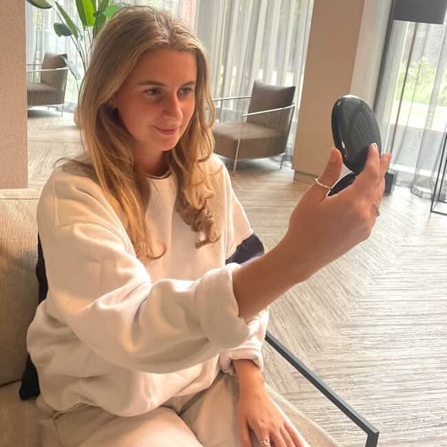 A young woman with long blonde hair, in a white sweater, sits on a sofa in a bright modern room and smiles as she looks into the Black Compact Mirror with Light.