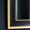 Close-up of the Hudson Black & Gold Full Length Mirror by Hollywood Mirrors, featuring a rectangular design with bevelled glass, sleek black frame, and gold metal border, reflecting part of a dark wall.