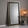 The Hollywood Mirrors Hudson Black & Gold Full Length Mirror with bevelled glass leans against a gray wall beside a round side table topped with a modern lamp featuring a geometric wire shade and a small stack of books.